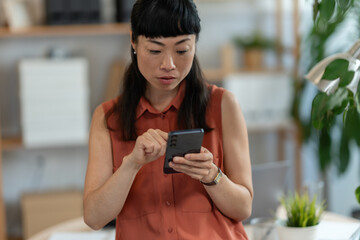Focused Asian small-business owner using her smartphone in a bright home office, checking important...