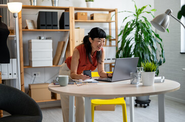 Fototapeta premium Asian woman small entrepreneur working in her cozy home office, standing at her desk while reviewing notes and using her laptop in a bright, modern workspace