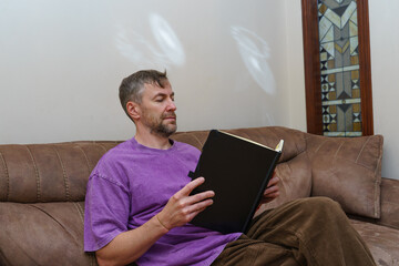 Casual man sitting on a sofa at home reading a book. Focused on learning and leisure