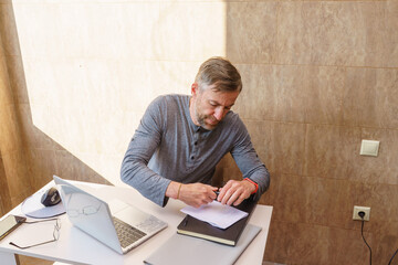 Focused middle-aged man writing on paper at his desk. Professional working remotely with a laptop