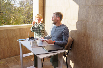 Playful son distracts father working from home. Man with laptop on balcony enjoys family time