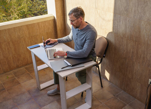 Middle-aged man working on a laptop at a desk on a sunny balcony. Remote work lifestyle