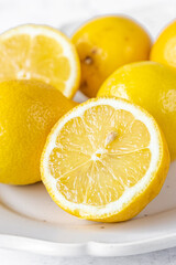 Bright fresh lemons and lemon halves arranged on a cream ceramic plate against a light marble background. Clean, vibrant food still life ideal for culinary, recipe, and citrus-themed designs.