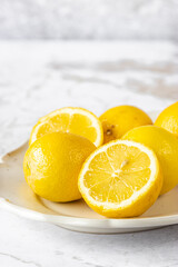 Bright fresh lemons and lemon halves arranged on a cream ceramic plate against a light marble background. Clean, vibrant food still life ideal for culinary, recipe, and citrus-themed designs.