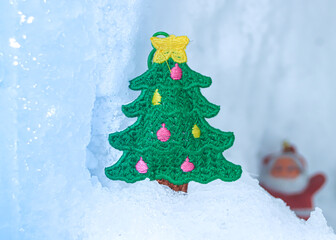 Knitted Christmas tree patch on a glacial ice base with blurred Santa doll in the soft background. Serene winter holiday scene with ample white space. Perfect for seasonal greetings.
