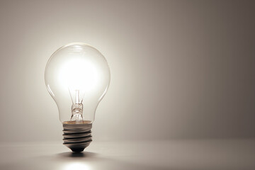A brightly glowing incandescent light bulb stands upright on a subtle gradient gray background. Clean, minimalist studio shot. Idea, innovation, solution concept. Copy space, place for signature.