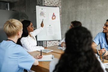 African american young woman chief physician making presentation in front of male and female international colleagues while team-building, pointing at presentation board, copy space