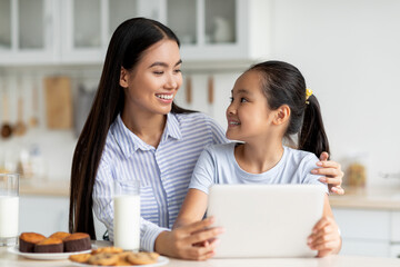 Loving mother and her daughter sit at the kitchen table, using a digital tablet to find new recipes. They share cookies and milk, enjoying a special moment together in a warm, inviting space.