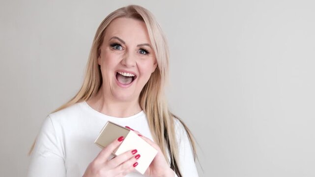 vibrant video set showing a happy forty five year old European woman holding and opening a gift box as she laughs dances and enjoys the moment with red nails white top long lashes.