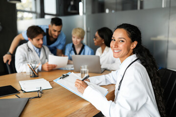 Middle eastern woman doctor smiling at camera while having morning session with colleagues, multiracial group of physicians professional doctors discussing medical cases, using modern technologies