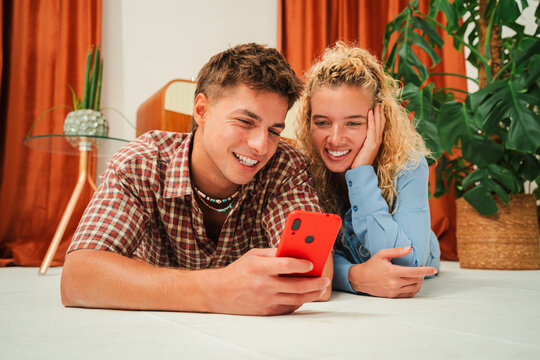 Two cheerful friends share a joyful moment as they excitedly explore an app on a smartphone while lying on the floor, showcasing their happy expressions and enthusiasm for the mobile device. - Powered by Adobe