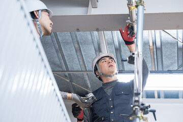 天井が近い高所の足場で単管パイプを固定するクランプなどの施工作業　鳶職や作業員のイメージ　レンチで締める2人　確認やチェック　ご安全に