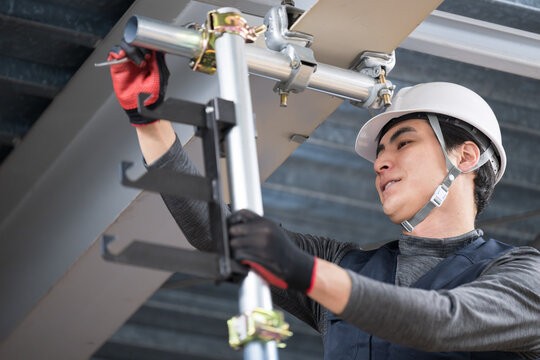 天井が近い高所の足場で単管パイプを固定するクランプなどの施工作業　鳶職や作業員のイメージ　レンチで締める若い男性　クローズアップ