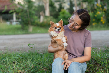 Happy Asian woman sitting on grass hugging and smiling at cute Chihuahua dog in park.