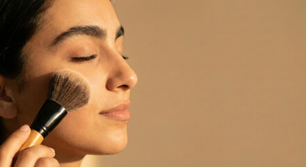 Side profile of beautiful young woman applying face powder with makeup brush in warm golden lighting
