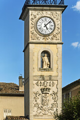 Ornate stone clock tower in Sisteron, France, featuring a circular clock face with Roman numerals and black hands showing 10:10. Below the clock, a statue of a seated figure is set in a niche,