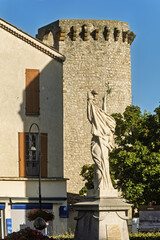 Stone statue of a robed figure holding an object aloft in a public square in Sisteron, France. Behind the statue stands a tall cylindrical tower with battlements, evoking medieval architecture.