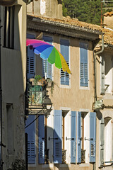 Charming street scene in Sisteron, France, with beige buildings featuring blue shutters and tiled roofs. A rainbow umbrella extends from a balcony with potted plants,
