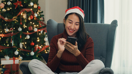 Happy Asia girl red santa hat new normal celebrate christmas party with smartphone send online greeting card to friend and family on wing chair in living room at house. Lifestyle technology concept.
