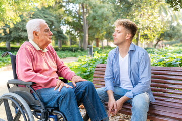 Grandson sitting on a bench talking with his grandfather in a wheelchair in a park