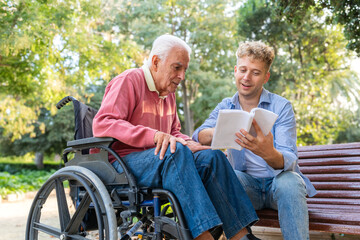 Grandson reading a book to his grandfather in a wheelchair while sitting on a bench at the park