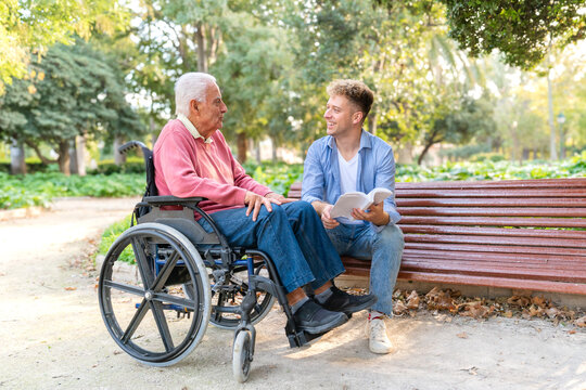 Young smiling man reading a book to his senior grandfather in a wheelchair at a park bench