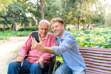 Smiling grandson sitting on bench with elderly man in wheelchair taking a selfie in a park