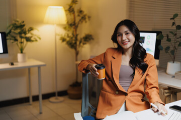 Smiling Asian businesswoman working late in office holding coffee