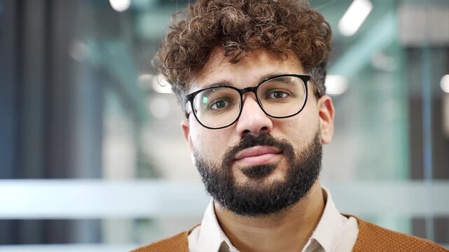 Portrait of serious handsome businessman standing at workplace in modern business office. Head shot of confident successful bearded male professional in glasses and sweater looking at camera. Close up