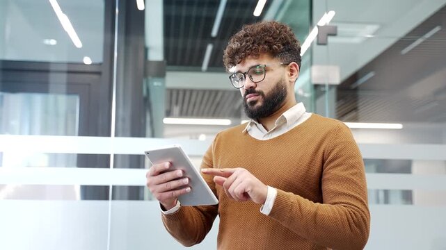Businessman is using a digital tablet while standing at workplace in a modern glass business office. Confident manager works on application, typing a message, chatting online with client, browsing web