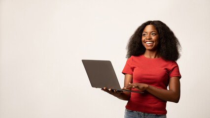 A happy young Black woman smiles while holding a laptop. She stands confidently, showcasing her...