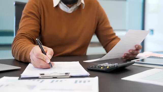 Close up. Man's hand fills out a form with a pen at a desk at a workplace in a business office. A businessman or financier is doing paperwork, writing, making a tax return, works with documents
