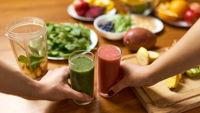 Hands holding two smoothies above wooden table with greens fruit and blender jar. Concept of wellness marketing, smoothie product visuals, nutrition planning guides, healthy eating tutorials.