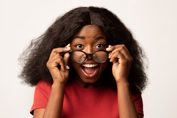 A joyful Black woman is taking off her glasses while looking at the camera, expressing surprise and...