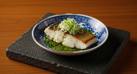 Grilled fish fillet with green onions and sauce in a decorated ceramic bowl on a dark stone slab, placed on a wood table