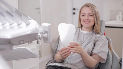 Caucasian female with blonde hair smiling in dental clinic holding mirror, white light background.