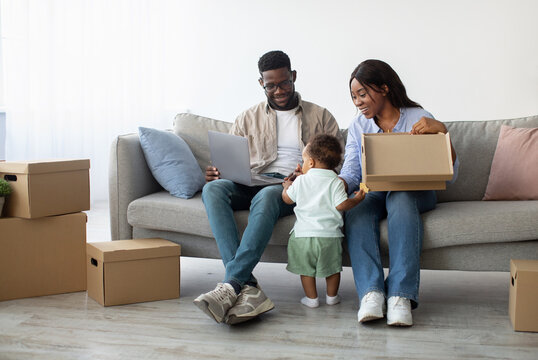 A joyful family of three is relaxing together on a sofa in their new home. They are using a computer while surrounded by cardboard boxes. Their child is exploring the boxes.
