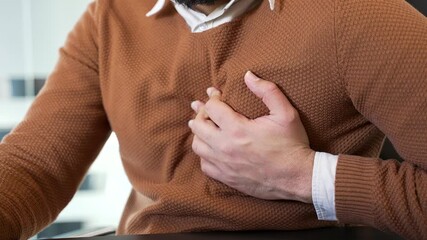 Close up. Businessman in casual sweater has a heart attack while sitting at a workplace in a business office. Male hands holding massaging chest. Sick man worker suffers from pain, needs medical help