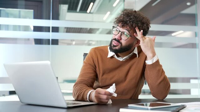 Sick businessman suffers from a cold and headache while sitting at a desk at a workplace in a business office. Unhealthy and tired male has symptoms of virus or flu. Unwell man has fever and feels bad