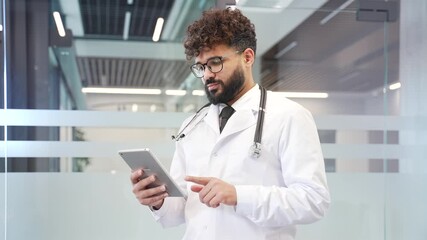 Doctor in white coat using digital tablet standing in modern hospital office. Bearded medical worker physician in glasses chats online with patient, browses information on web or works in application - Powered by Adobe