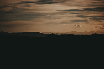 Mountains silhouette under a cloudy sky at sunset. The sun sets behind the mountain range, casting a warm glow on the clouds, creating a serene and peaceful atmosphere. Moody landscape in twilight. 