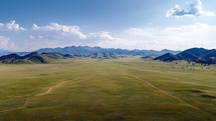Vast green prairie landscape with rolling hills, a dirt track, and a mountain range in the distance under a bright, cloudy sky.