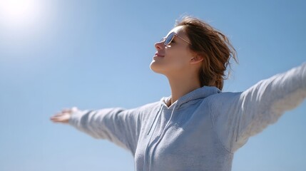 A joyful young woman in sunglasses and a grey hoodie opens her arms wide to the clear blue sky feeling the warmth of the bright sun and embracing a sense of freedom and peace