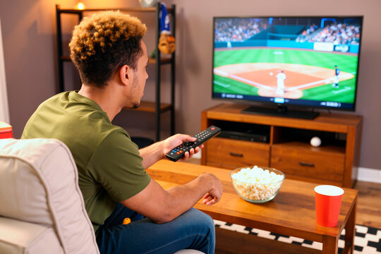 Young adult Black man sitting on sofa holding remote control watching baseball game on television with bowl of popcorn and drink on table in living room setting