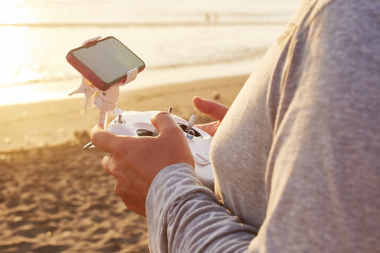 Close up of unrecognizable person operating drone remote controller with mounted smartphone on sandy beach, hands gripping device while focusing on piloting, ocean visible in background