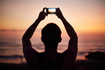 Man holding smartphone above head capturing sunset over ocean, standing outdoors with arms raised, silhouetted against colorful sky during evening