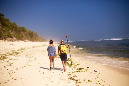 Young woman and man walking along sandy beach near ocean, man carrying backpack and holding walking stick, both moving away from camera toward distant shoreline