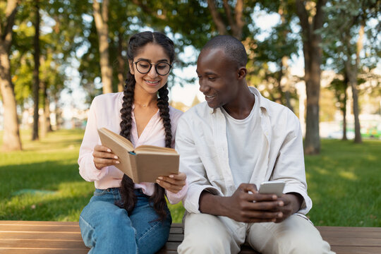 Two young people share a peaceful moment in the park. One reads a book, while the other checks their smartphone, both smiling under the sun surrounded by trees.
