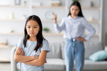 In a living room, a daughter stands with crossed arms, ignoring her mother who appears frustrated. The girl shows her displeasure, emphasizing the family conflict between them.