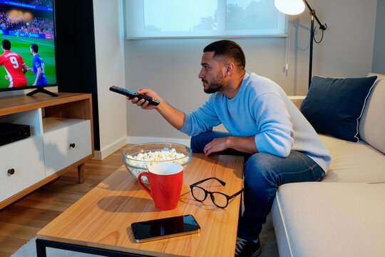 Young adult Caucasian man sitting on sofa watching soccer match on television holding remote control with focused expression, popcorn and coffee mug on table in living room